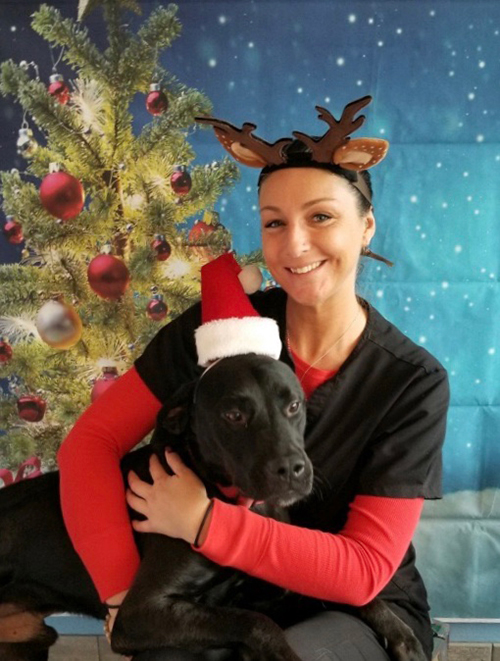 A pretty woman wearing black scrub top and a red shirt underneath with a reindeer headband cuddles a black dog, who is wearing a red and white Santa hat. Behind them is a Christmas tree background.