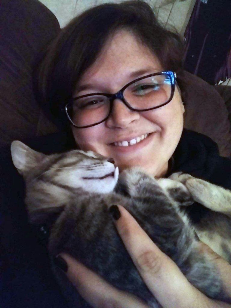 A woman with brown hair and wearing glasses holds a grey striped cat, who looks like it may be sleeping and has its paws curled up near its face.